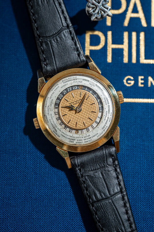 Patek Philippe World Time reference 2523 in yellow gold, salmon guilloche centre, silvered city ring and black alligator strap, photographed on blue fabric beneath a Patek Philippe Geneve sign