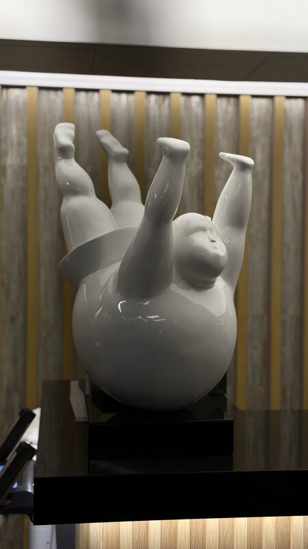 Glossy white sculpture of a rounded figure lying back on a sphere with limbs in the air, in a lobby with gold vertical blinds