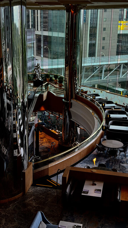 Looking down through glass into a circular multi-level atrium with lounge seating, Hong Kong buildings behind