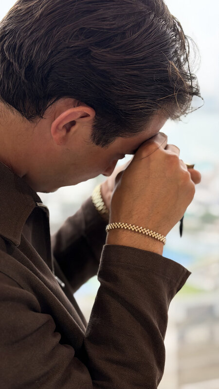 A man in a brown suit with a gold bracelet watch, head bowed, holding a loupe to his eye