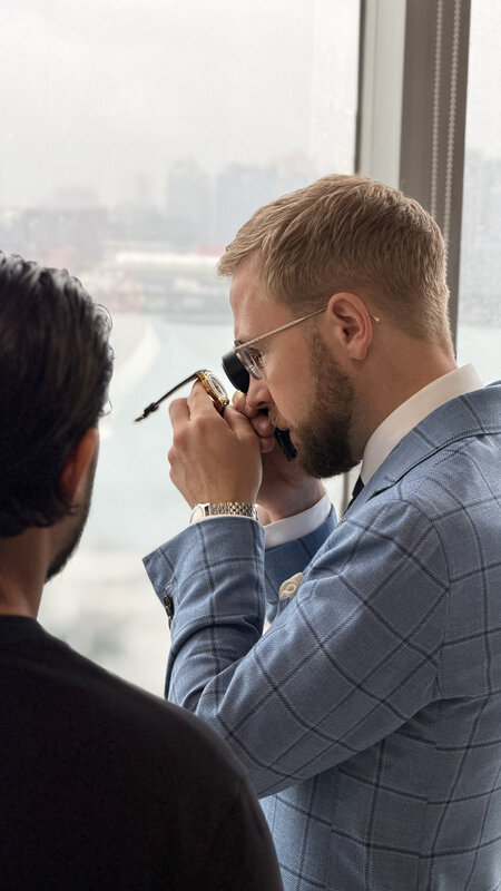 A bearded man in a blue windowpane jacket examining a watch through a loupe by the window, another man in the foreground