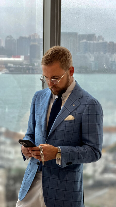 A man in a blue windowpane jacket and glasses looking at his phone by the window, the harbour behind him