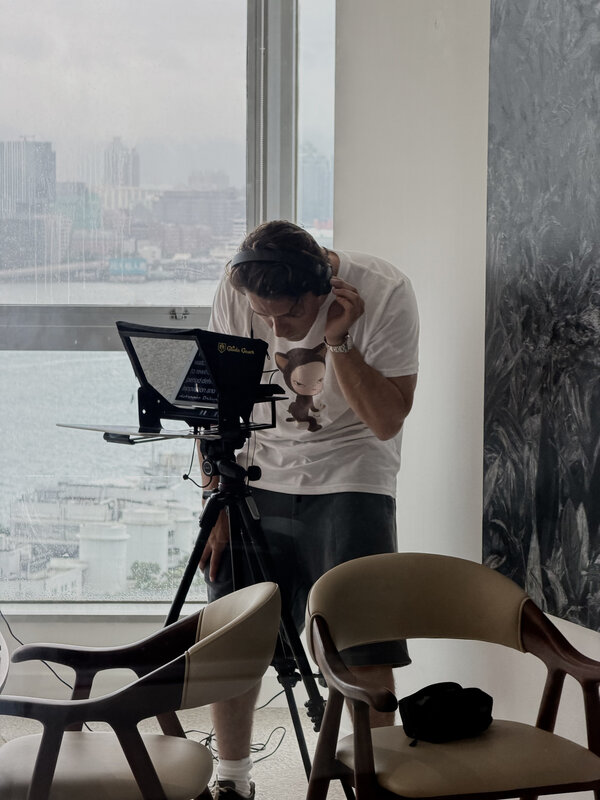 A Glide Gear teleprompter and microphone on a tripod, script text on the screen, a man pulling on headphones behind it, harbour windows beyond