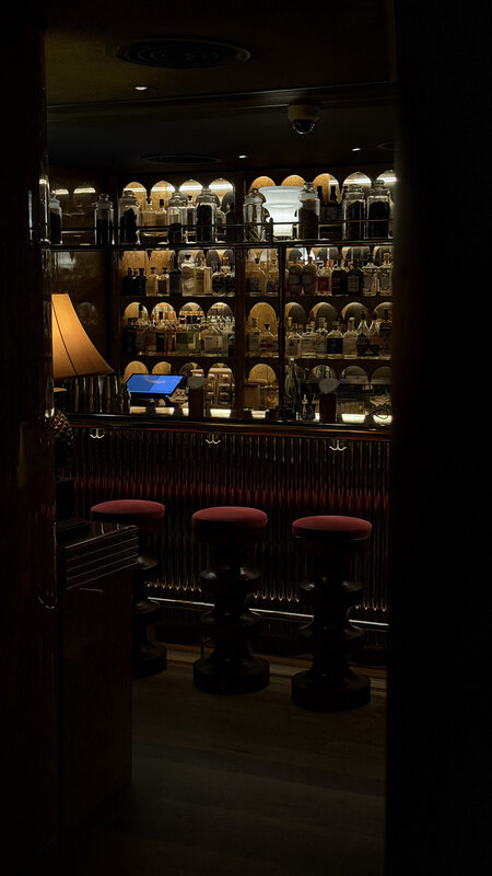 A dark hotel bar in Hong Kong with backlit shelves of bottles above the counter and low red stools