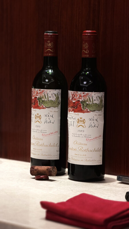 Two bottles of Château Mouton Rothschild 1989 on a marble counter against dark wood, a cork and a folded red napkin beside them