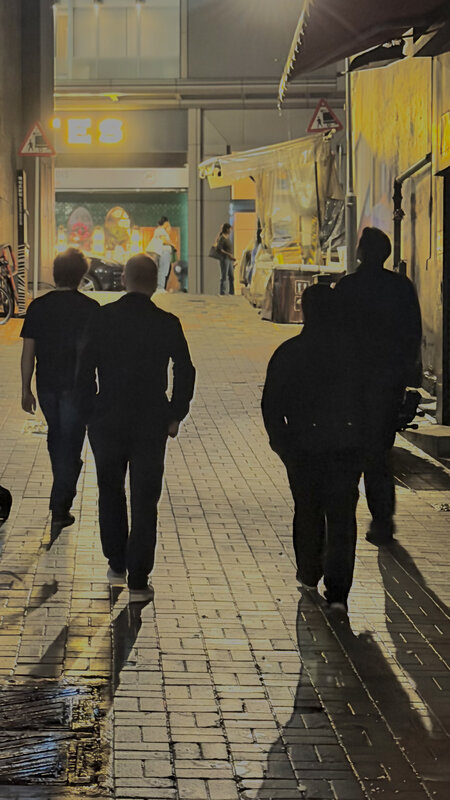 Silhouettes of four people walking down a narrow Hong Kong alley at night, lit yellow signage on both sides