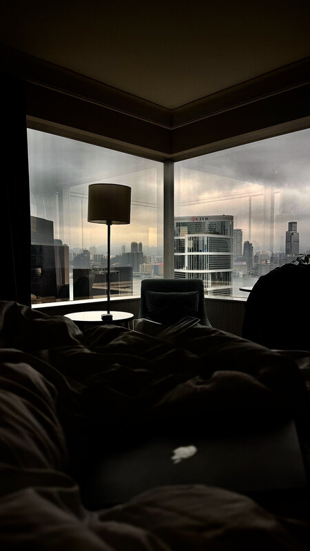 Hotel room in low light, an unmade bed in the foreground and a grey, overcast Hong Kong skyline through the window