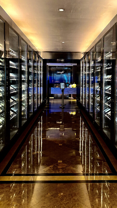 A corridor of lit glass wine cabinets reflected in a polished marble floor, a cafe sign at the far end