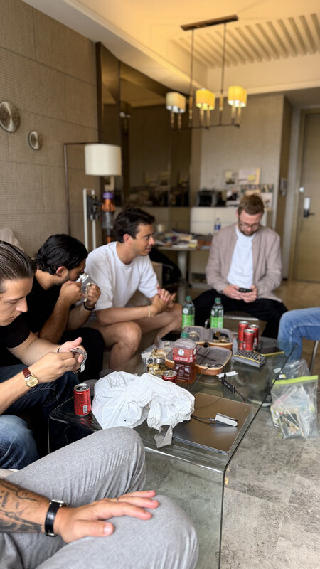 Five men around a low glass table in a Hong Kong apartment, daylight from the window behind, a cigar box, cans and cleaning cloths spread across the glass