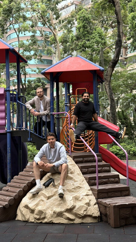 Three men on a playground climbing frame in a Hong Kong park, tower blocks and trees rising behind