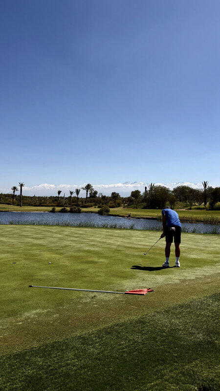 Wide Royal Palm Golf fairway — water hazard, a player in blue putting on the green, Atlas Mountains in the distance
