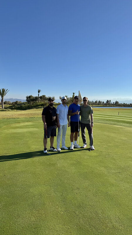 Four friends standing together on a Royal Palm Golf green, Atlas Mountains in the distance, clear sky