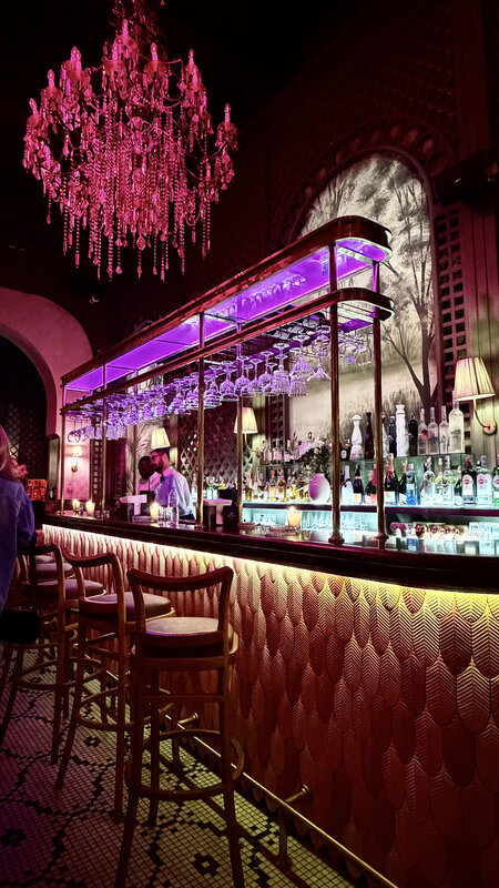 Bar interior at night with a large pink crystal chandelier, full liquor wall, leather barstools, upscale Marrakesh setting