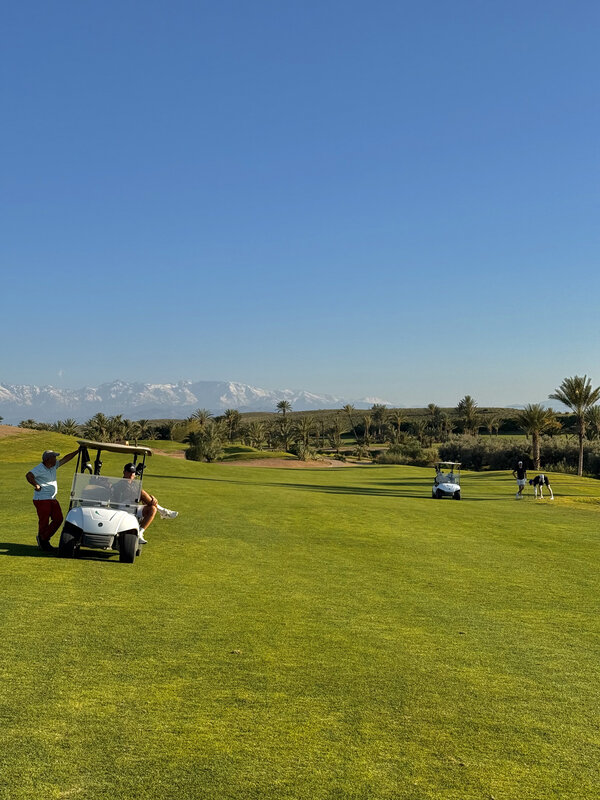 Wide Royal Palm Golf view — caddies and buggies in the foreground, snow-capped Atlas Mountains in the distance