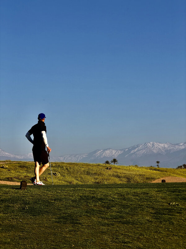 Jimmy in silhouette standing on the tee box, snow-capped Atlas Mountains in the distance, late afternoon light