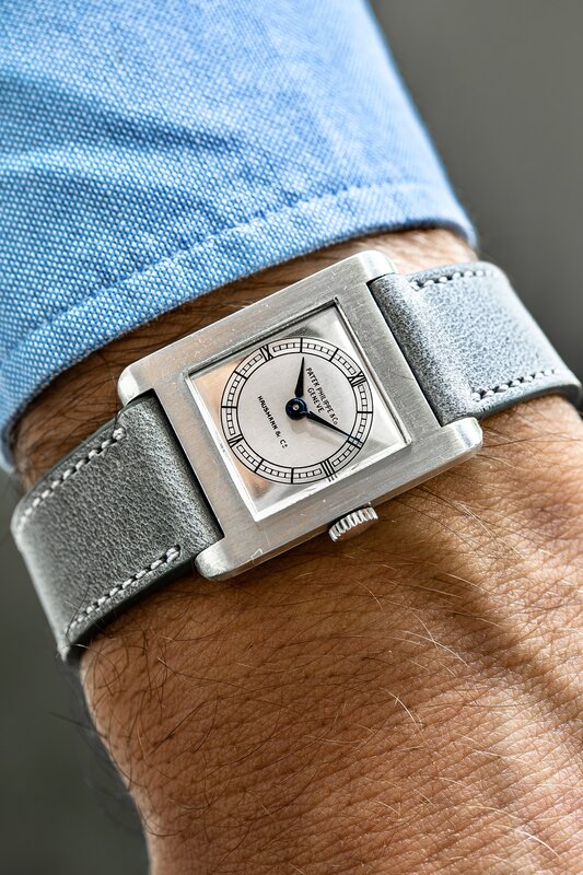 Silver rectangular Reverso-style dress watch on wrist with grey leather strap, blue suit sleeve