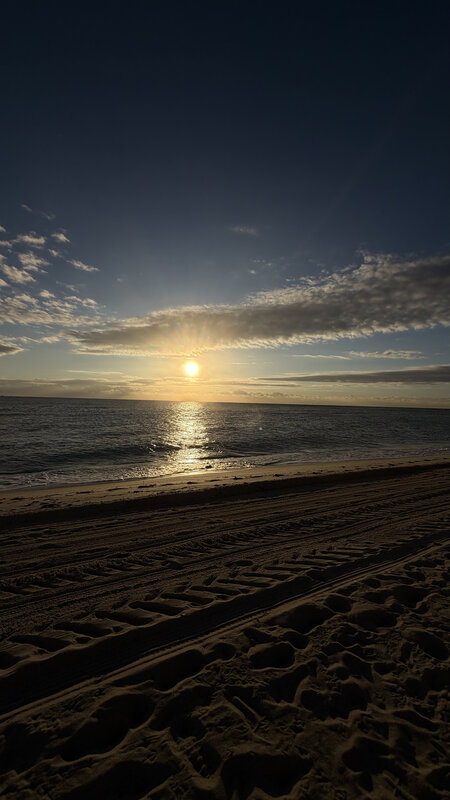 Wide view of Miami Beach at sunrise — sand tracks, ocean, distant condo towers, sun behind clouds