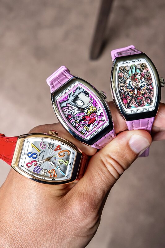Three Franck Muller Crazy Hours watches held in one hand, a red-strapped colour-numeral piece and two pastel-strapped artist-edition dials with hand-painted graffiti motifs