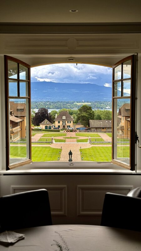 Open window framing the Watchland estate, formal garden parterre, ochre manor houses, and Lake Geneva with the mountains beyond