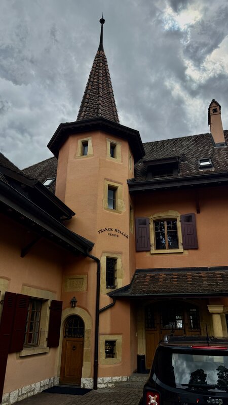 The historic Watchland château in Genthod, an ochre tower engraved Franck Muller Genève and dated 1905, beneath a dramatic cloudy sky