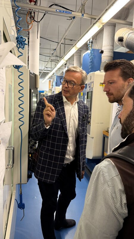 Nick Rudaz explaining a CNC machine on the manufacture floor, gesturing as two visitors look on amid the workshop equipment