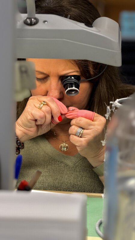 A watchmaker at the bench wearing a loupe headband, inspecting a tiny component held in pink finger cots