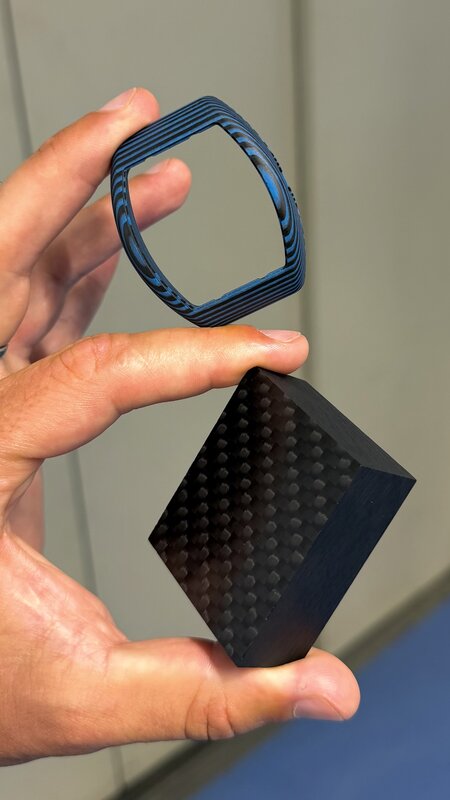 A hand holding two case materials side by side, a blue layered-metal tonneau frame and a black carbon-fibre block