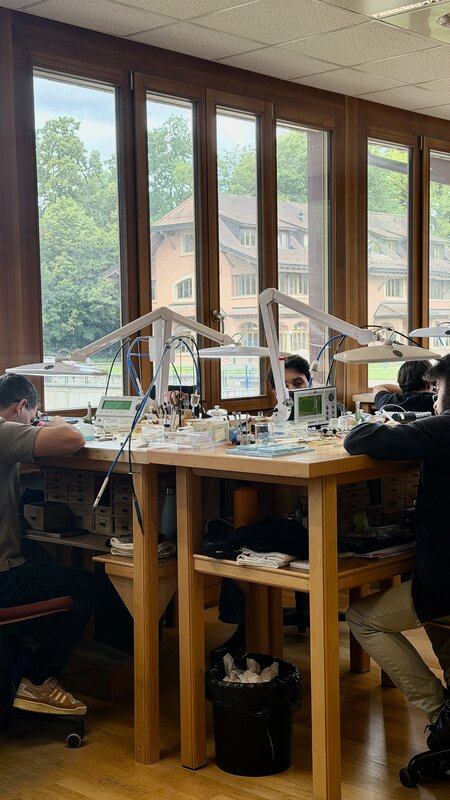 Watchmakers working at lamp-lit benches by tall windows that overlook the Watchland grounds and its ochre manor