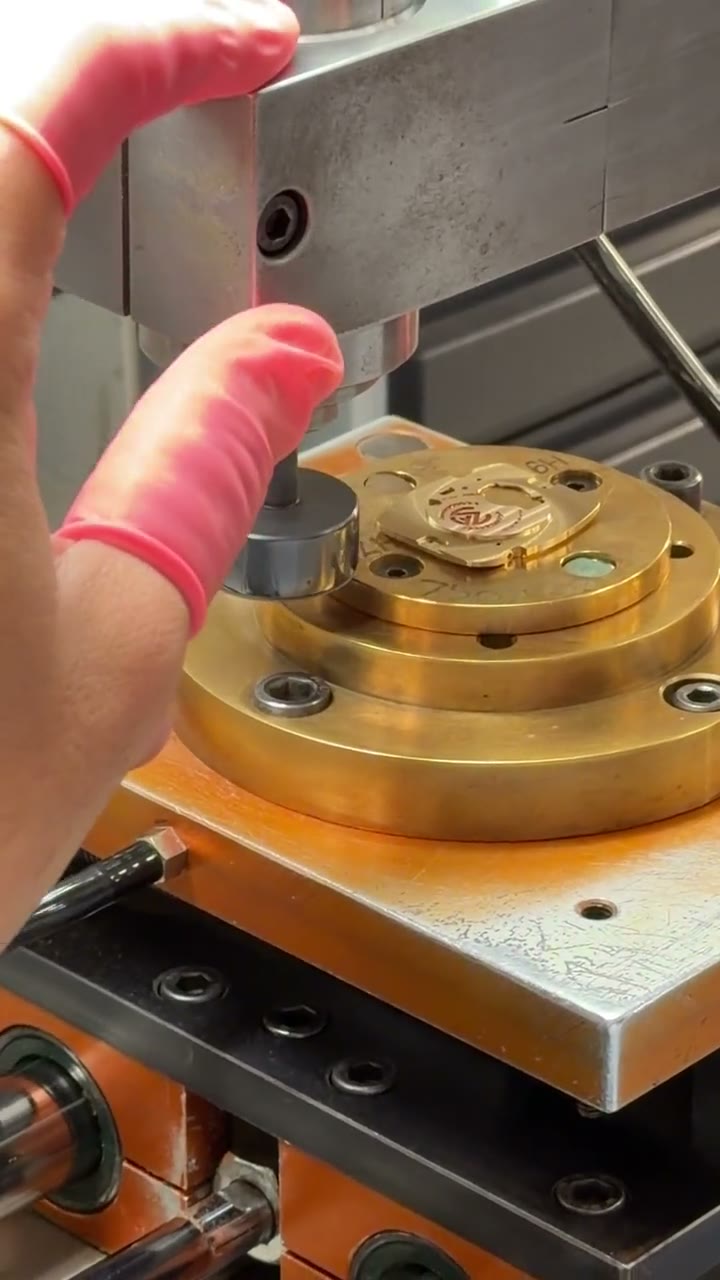 Hands in finger cots guiding a gilt Franck Muller movement plate on a cross-slide table beneath a rotating finishing tool at the manufacture
